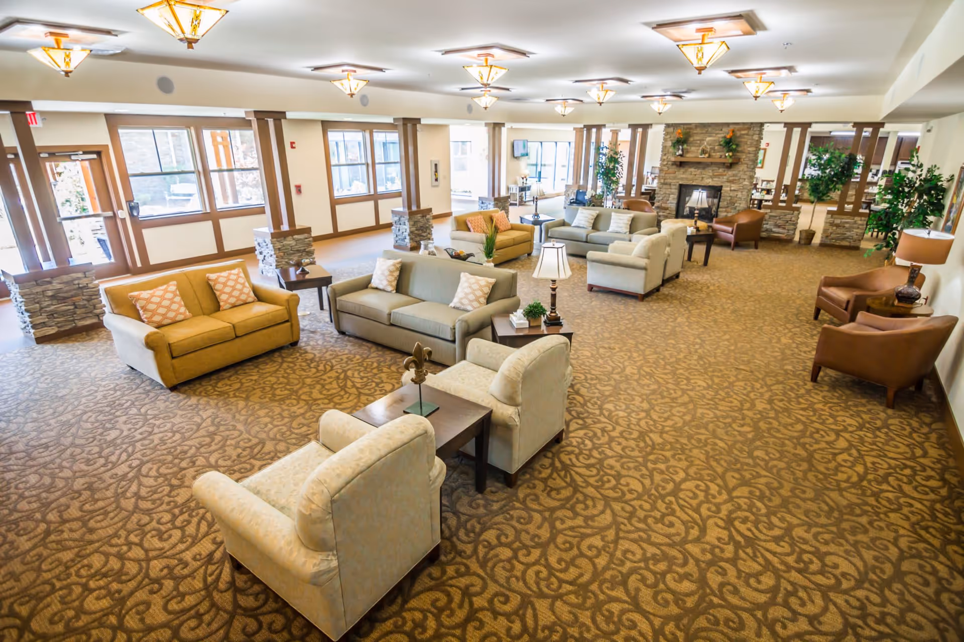 A spacious and well-lit senior living facility common area with multiple seating arrangements including sofas and armchairs. The room features large windows, decorative pillars with stone bases, patterned carpet, and a stone fireplace with plants and decorations on the mantel. Several lamps and ceiling lights provide warm lighting throughout the space.