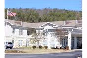 Exterior view of a single-story senior living facility building with an American flag on a flagpole in front, surrounded by a driveway and some landscaping with trees and bushes. The building has multiple windows and a covered entrance.