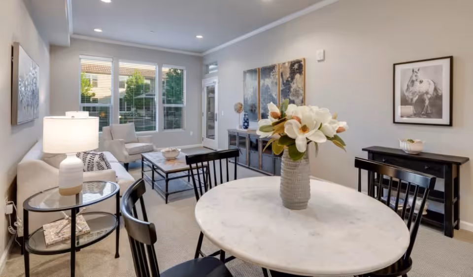 A bright and cozy living room area with a round white marble table in the foreground, decorated with a vase of white flowers. The room features a beige sofa, an armchair, a glass-top side table with a lamp, a coffee table, and a console table against the wall. Large windows with white blinds allow natural light to fill the space. The walls are adorned with framed artwork, including a horse picture and a triptych painting.
