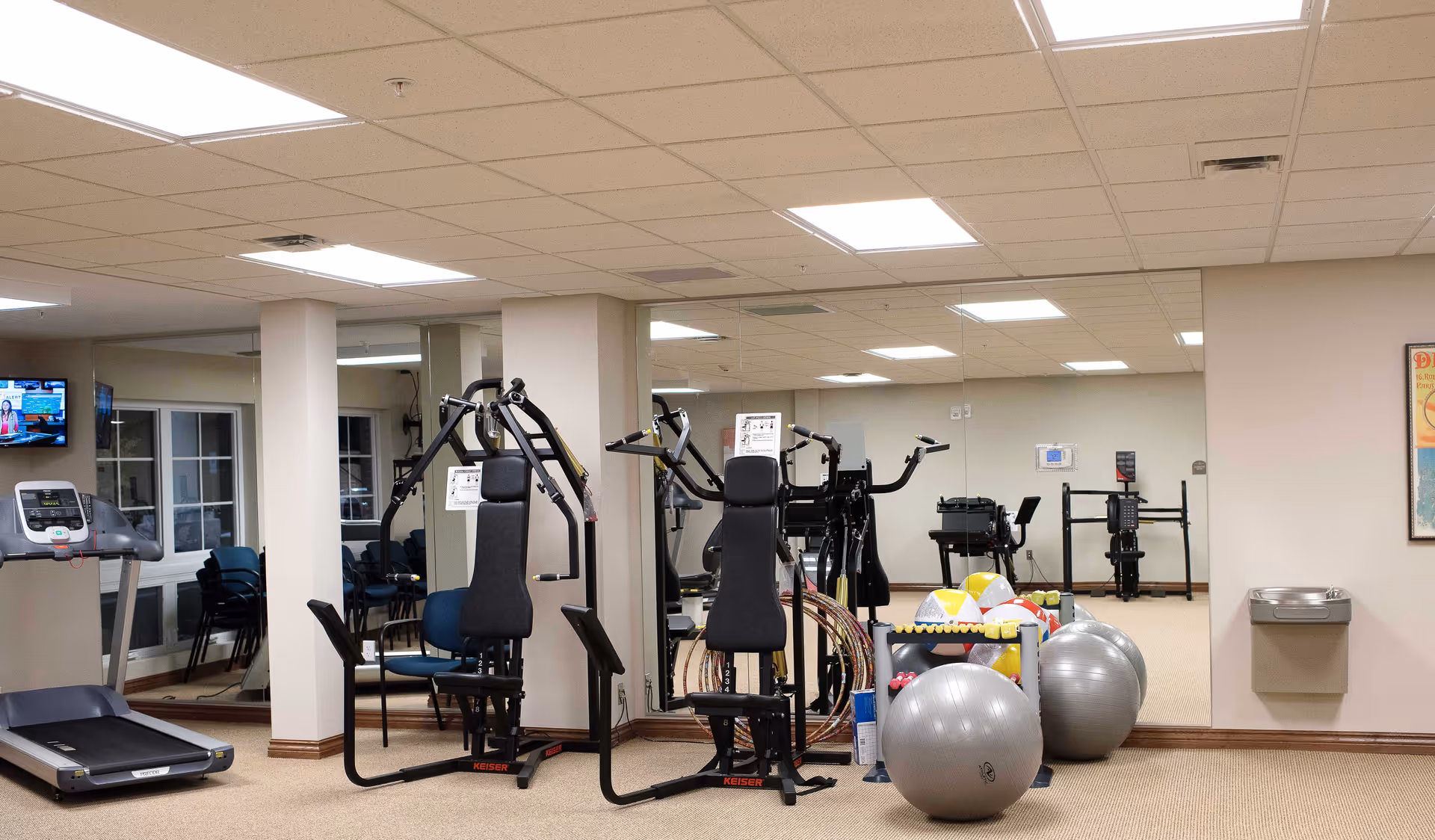 Interior view of a fitness room in a senior living facility with exercise equipment including a treadmill, weight machines, exercise balls, and hula hoops. The room has beige walls, a carpeted floor, a water fountain, and large mirrors on one wall reflecting the equipment.