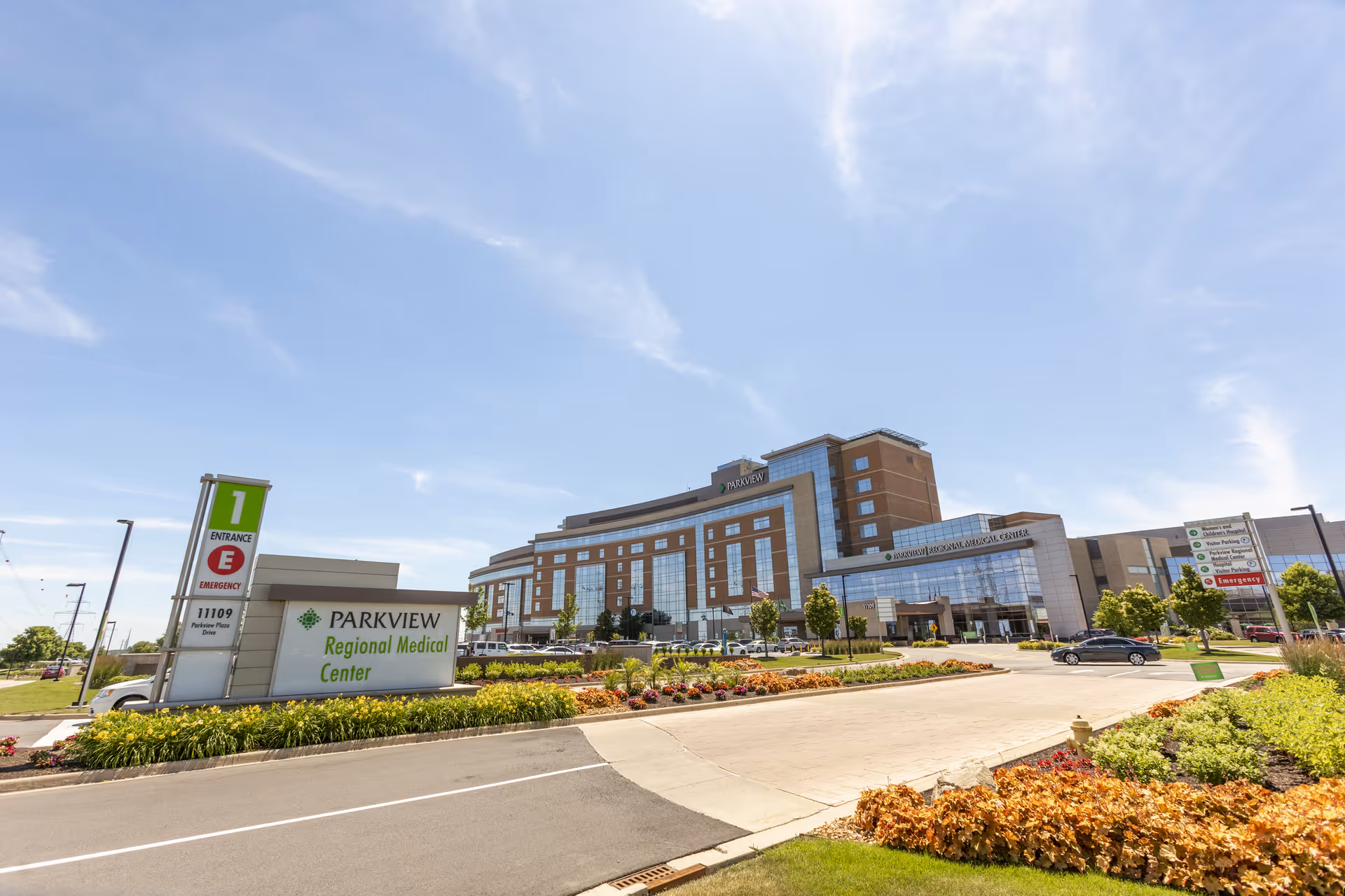 Exterior view of Parkview Regional Medical Center on a sunny day with clear blue sky, showing the main hospital building, entrance signs, landscaped gardens, and a driveway with cars.