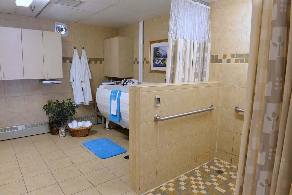 A spacious bathroom with beige tiled walls and floor. The room features a walk-in shower with a grab bar and a patterned shower curtain. There is a white bathtub with a blue towel hanging over the side, two white bathrobes hanging on hooks, a basket with rolled white towels, a blue bath mat, and a potted plant. Cabinets are mounted on the wall for storage, and a framed picture hangs on the wall above the bathtub.
