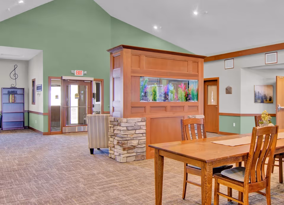 Communal dining and lounge area with a wooden table and chairs, a built-in aquarium divider, and an entryway.