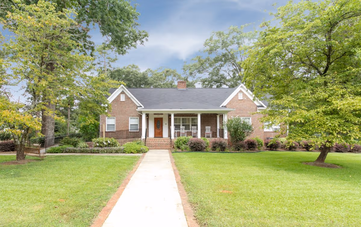 A single-story brick house with a front porch featuring rocking chairs, surrounded by green grass, trees, and shrubs under a partly cloudy sky.