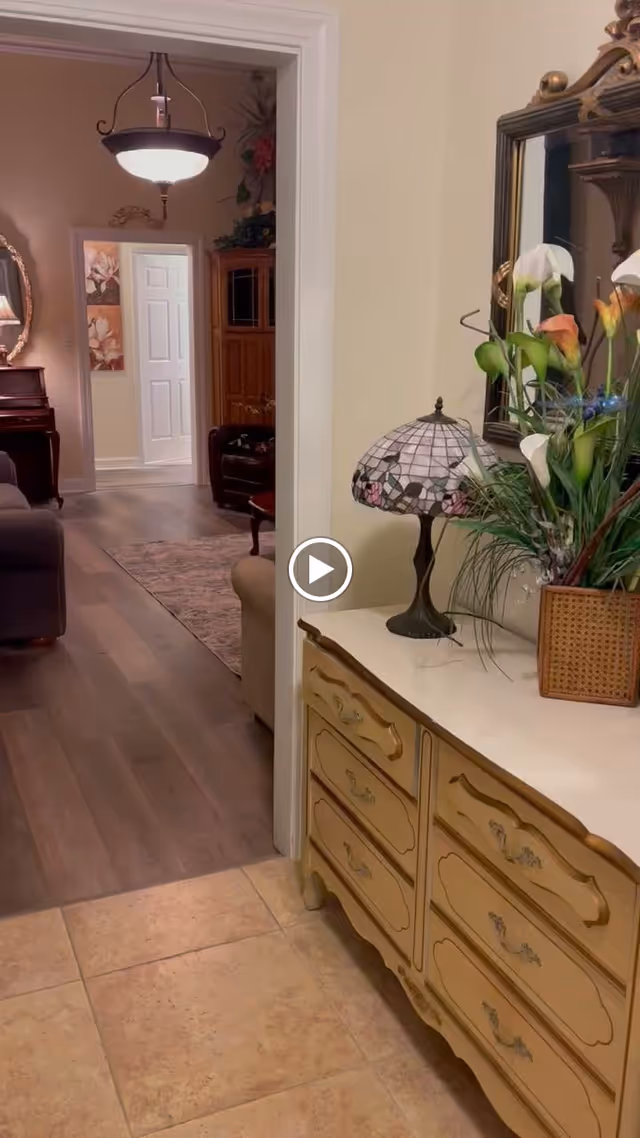 Hallway interior with a vintage dresser topped by a stained-glass lamp and floral arrangement, opening into a furnished living room.