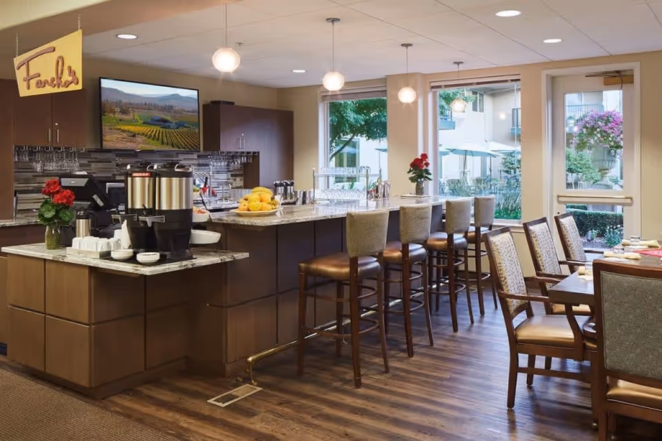 Interior view of a dining area in a senior living facility with a counter featuring coffee dispensers, bowls, and a fruit plate. There are high chairs along the counter and dining tables with chairs nearby. Large windows and a glass door provide a view of an outdoor patio with umbrellas and greenery.