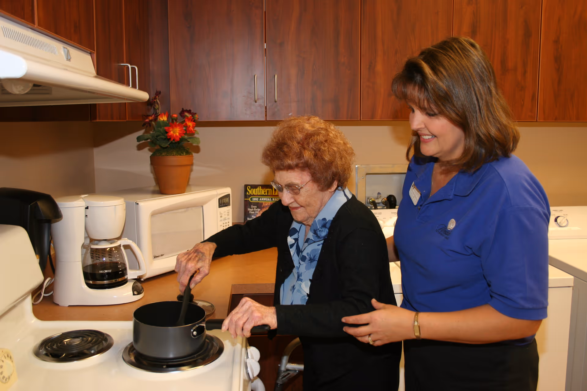 An elderly woman stirs a pot on a stove while a caregiver stands beside her in a kitchen area.