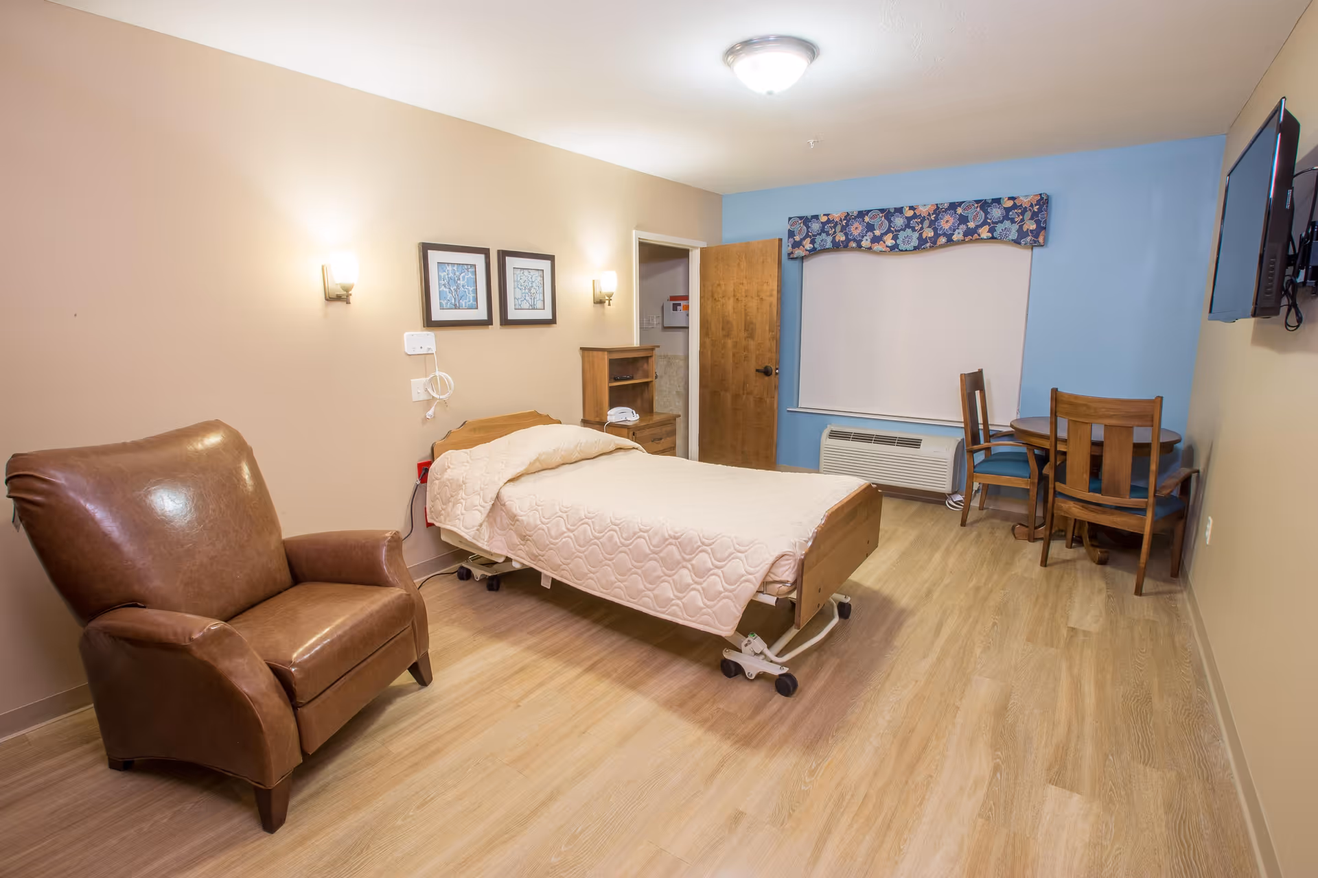 A clean and well-lit bedroom in a senior living facility featuring a single hospital-style bed with a beige quilt, a brown leather armchair, a small wooden table with three chairs near a window with a floral valance, a wall-mounted TV, and light wood flooring.