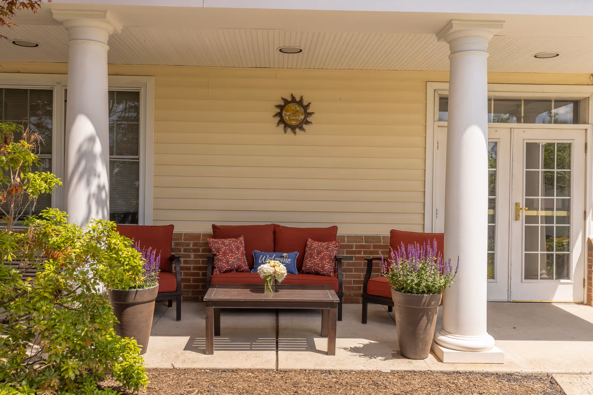 Outdoor seating area with a red cushioned sofa and two matching chairs around a wooden coffee table with a small flower arrangement. The seating area is on a porch with two white columns and a beige siding wall with a decorative sun hanging above the sofa. Two large potted plants with purple flowers flank the seating area. A set of white double doors with glass panes is visible on the right side.