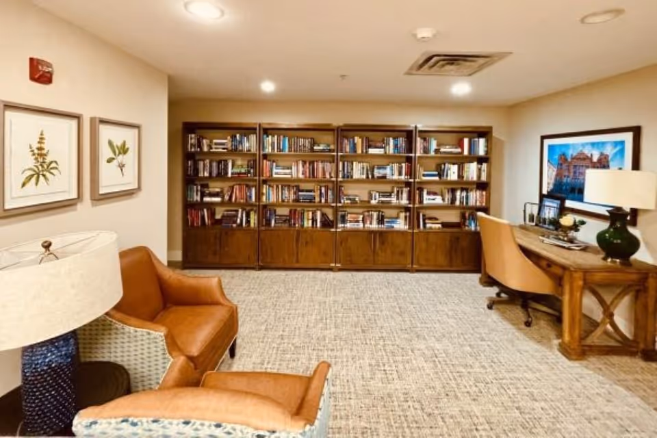 A cozy reading and work area in a senior living facility featuring a large wooden bookshelf filled with books against the back wall, two brown leather armchairs, a wooden desk with a beige office chair, a green table lamp, framed botanical prints on the wall, and a framed picture above the desk.
