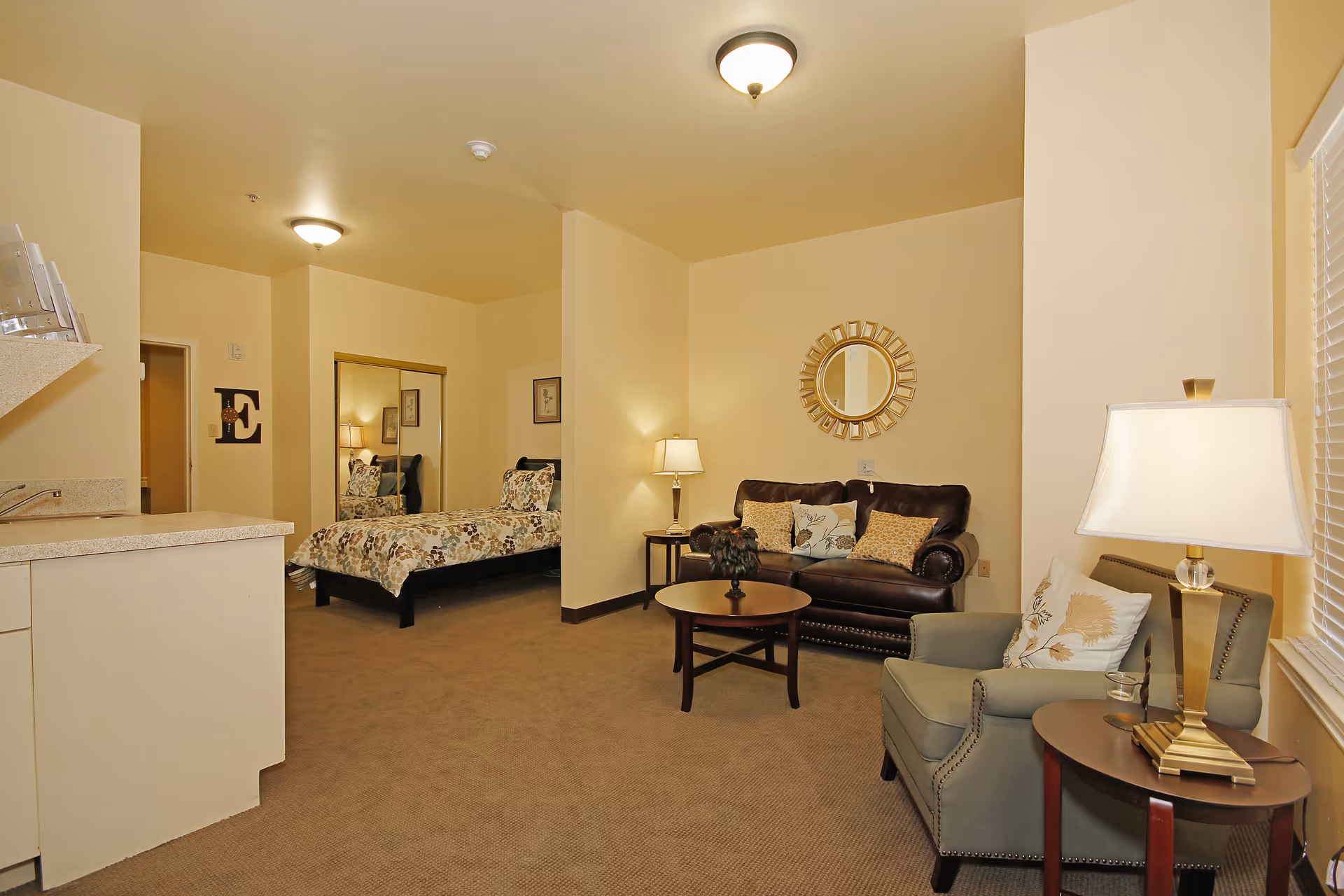 A cozy living space in a senior living facility featuring a small kitchenette on the left, a sitting area with a leather loveseat, an armchair with a side table and lamp, and a round coffee table. In the background, there is a bed with floral bedding and a mirrored closet door. The walls are painted beige, and the room is softly lit with ceiling lights and lamps.