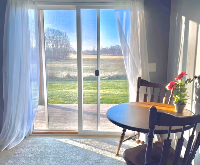 Sunlit dining nook with a round wooden table, chairs and a potted flower in front of a sliding glass door overlooking a grassy field.