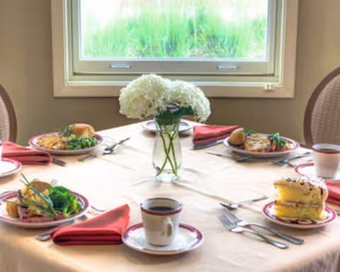 Round dining table set for a meal with plates of food, coffee cups, red napkins and a vase of white flowers in front of a window.