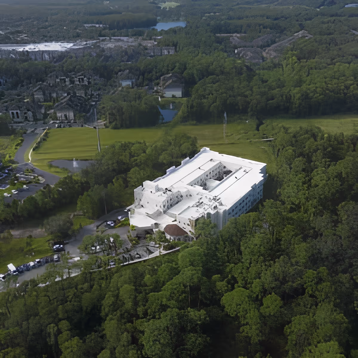 Aerial view of a large white building surrounded by dense green forest and open grassy areas, with a few parked cars visible near the building.