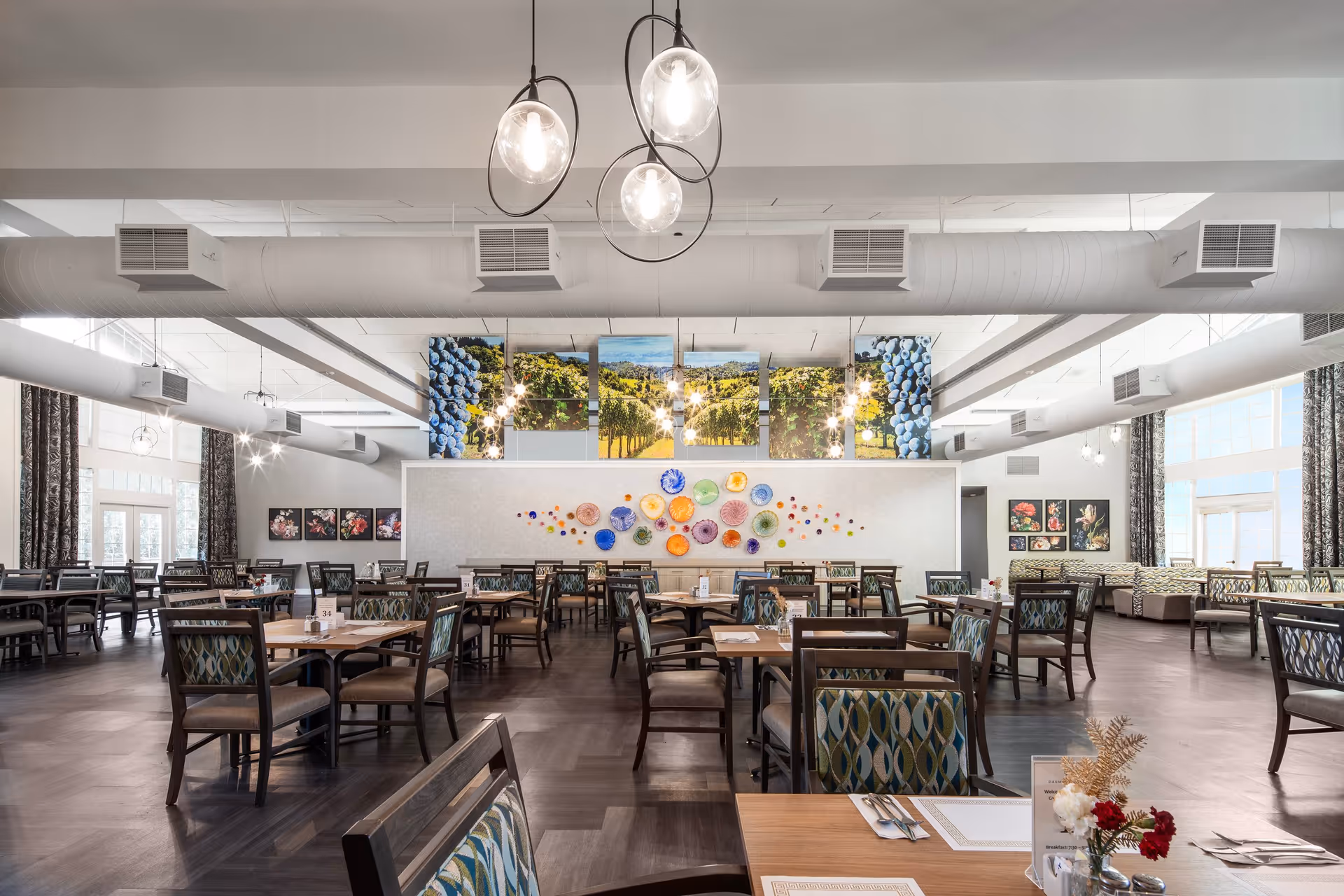 A spacious and well-lit dining room with multiple wooden tables and chairs arranged neatly. The room features large windows with patterned curtains, modern hanging light fixtures, and colorful wall art including a series of vineyard photos and decorative plates. The floor is dark wood, and the overall atmosphere is bright and inviting.