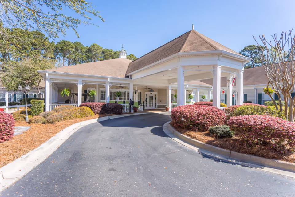 Front exterior view of a senior living facility named Oaks At Salem Road, featuring a covered driveway entrance with white columns, well-maintained landscaping with bushes and trees, and a clear blue sky.