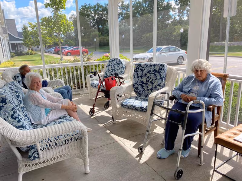 Three elderly individuals sitting on a covered porch with white wicker furniture that has blue patterned cushions. Two of them are seated on a wicker loveseat and chair, while the third is seated on a wooden chair using a walker. There are trees, parked cars, and a building visible outside the porch.