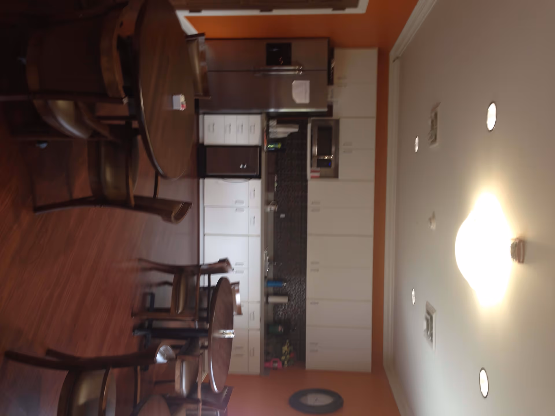 Interior view of a communal kitchen and dining area with several round wooden tables and chairs. The kitchen features white cabinets, a stainless steel refrigerator, microwave, and a dark backsplash. The walls are painted orange and the ceiling has recessed lighting and a central light fixture.