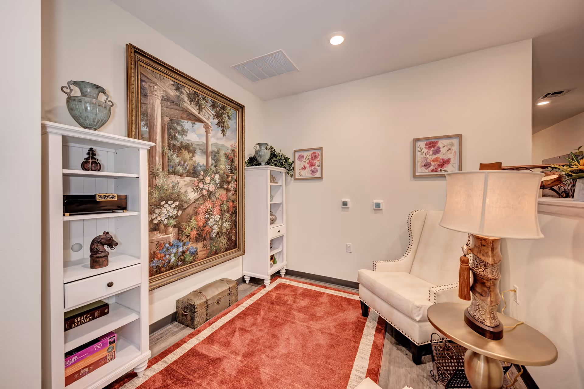 A cozy sitting area with a white upholstered armchair next to a small round table holding a decorative lamp. The room features a large framed painting of a garden scene with columns and flowers on the wall, two white bookshelves with decorative items and books, a red area rug with a white border on the floor, and two floral framed pictures on the wall.