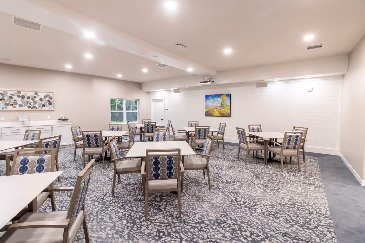 A bright and spacious dining room with multiple square and round tables surrounded by wooden chairs with patterned blue and beige cushions. The room has a patterned carpet, white walls, recessed ceiling lights, a window, and two pieces of artwork on the walls.