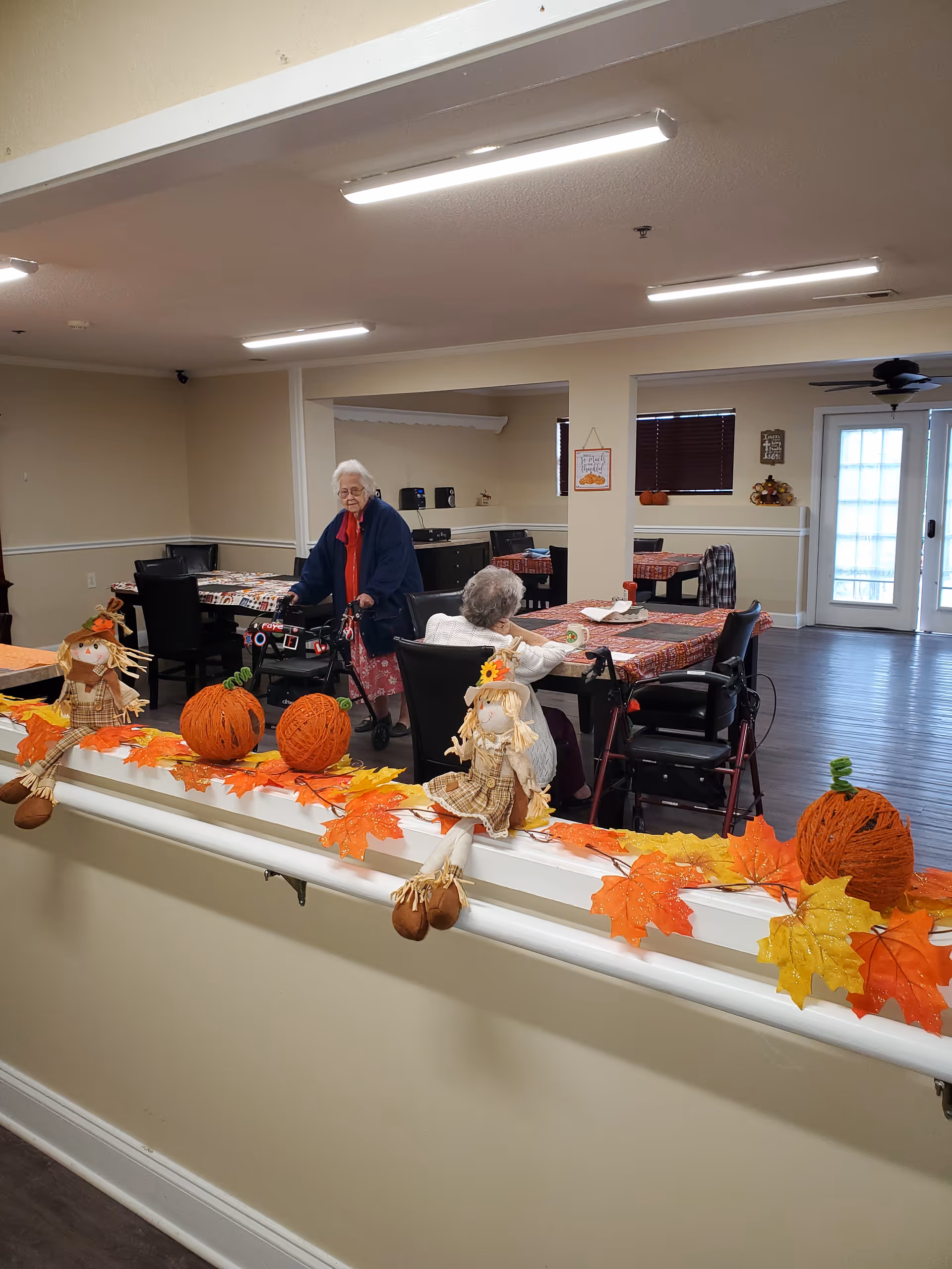 A senior living facility common area decorated with autumn-themed decorations including artificial pumpkins, scarecrows, and fall leaves on a white railing. Two elderly women are present, one standing with a walker and the other sitting at a table covered with a red checkered tablecloth. The room has beige walls, dark wood flooring, and fluorescent ceiling lights.