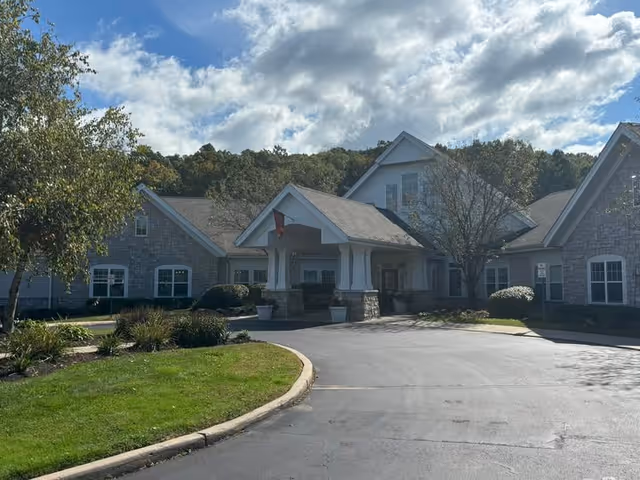 Exterior view of a single-story building with stone and siding facade, a covered entrance with columns, surrounded by landscaped greenery and trees under a partly cloudy sky.