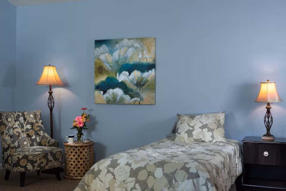 A decorated assisted-living bedroom with a single bed in floral bedding, a patterned armchair, two lamps, a side table with flowers, and framed artwork on a blue wall.