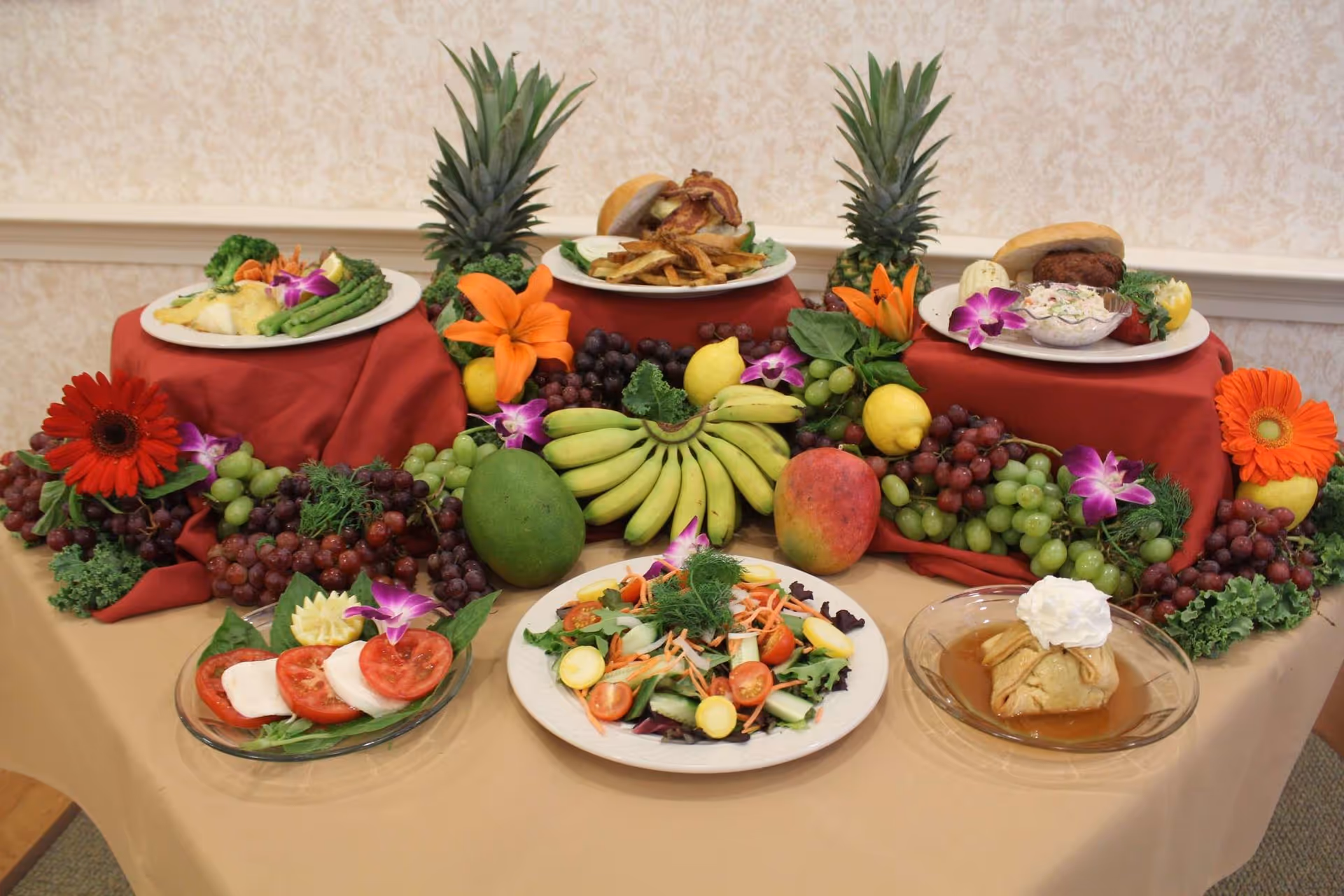 A table set with a variety of plated foods including a salad, a tomato and mozzarella dish, a dessert with whipped cream, and three elevated plates featuring a sandwich with fries, a burger with coleslaw, and a fish dish with asparagus. The table is decorated with an assortment of fresh fruits such as bananas, grapes, mango, and lemons, along with colorful flowers and greenery.