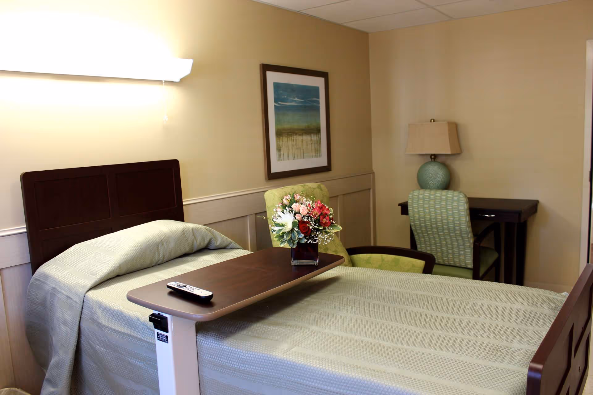 A tidy single bedroom in a senior facility with a made bed, overbed table holding a remote and a vase of flowers, an armchair, desk, and lamp.