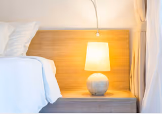 A bedside table with a round lamp emitting warm light, next to a bed with white bedding and pillows, against a wooden headboard and light-colored curtains.