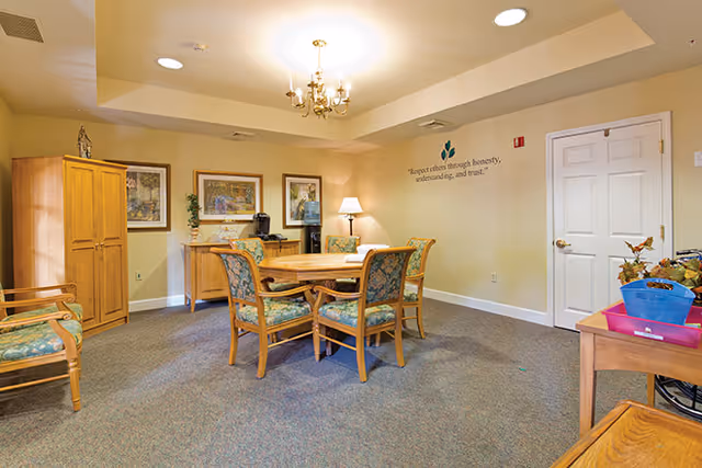 A cozy common area in a senior living facility featuring a wooden table with four cushioned chairs around it. The room has beige walls, carpeted floor, a wooden cabinet, framed artwork, a lamp on a side table, and a water cooler. A motivational quote is displayed on the wall, and there is a closed white door on the right side.