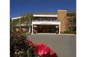 Front exterior view of Milford Health Care Center building with a driveway, trees, and blooming flowers in the foreground under a clear blue sky.