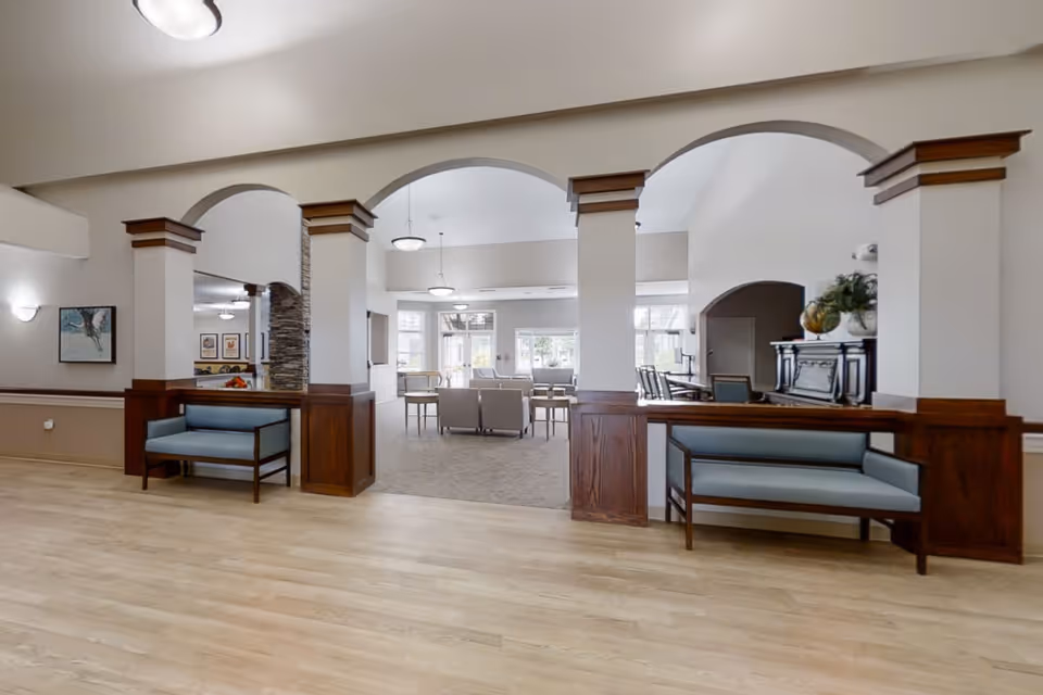 A spacious and well-lit interior common area with light wood flooring and white walls accented by wooden trim. The room features three large arches supported by columns with wooden paneling at the base. Two blue cushioned benches are placed against the columns. Beyond the arches, there is a carpeted seating area with armchairs and tables near large windows letting in natural light. Pendant lights hang from the ceiling, and a piano with decorative plants is visible on the right side.