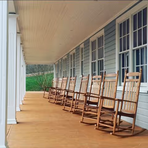 A long wooden porch with a row of wooden rocking chairs lined up against the light blue exterior wall of a building, with white framed windows and white columns supporting the porch roof.