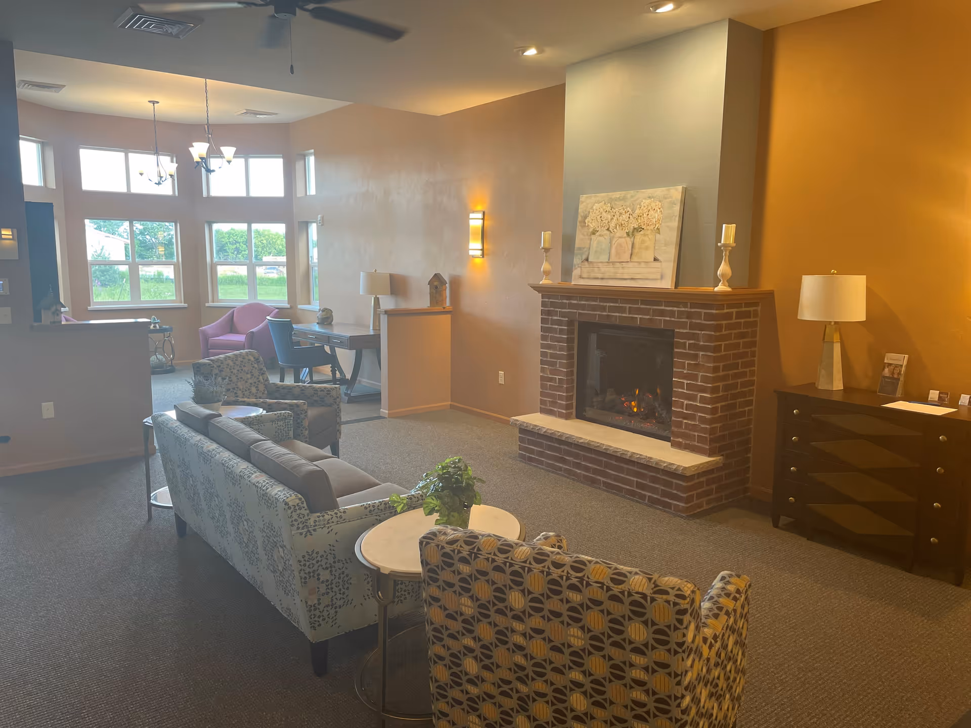 Cozy senior living room with sofas and armchairs arranged around a brick fireplace and large windows letting in daylight.
