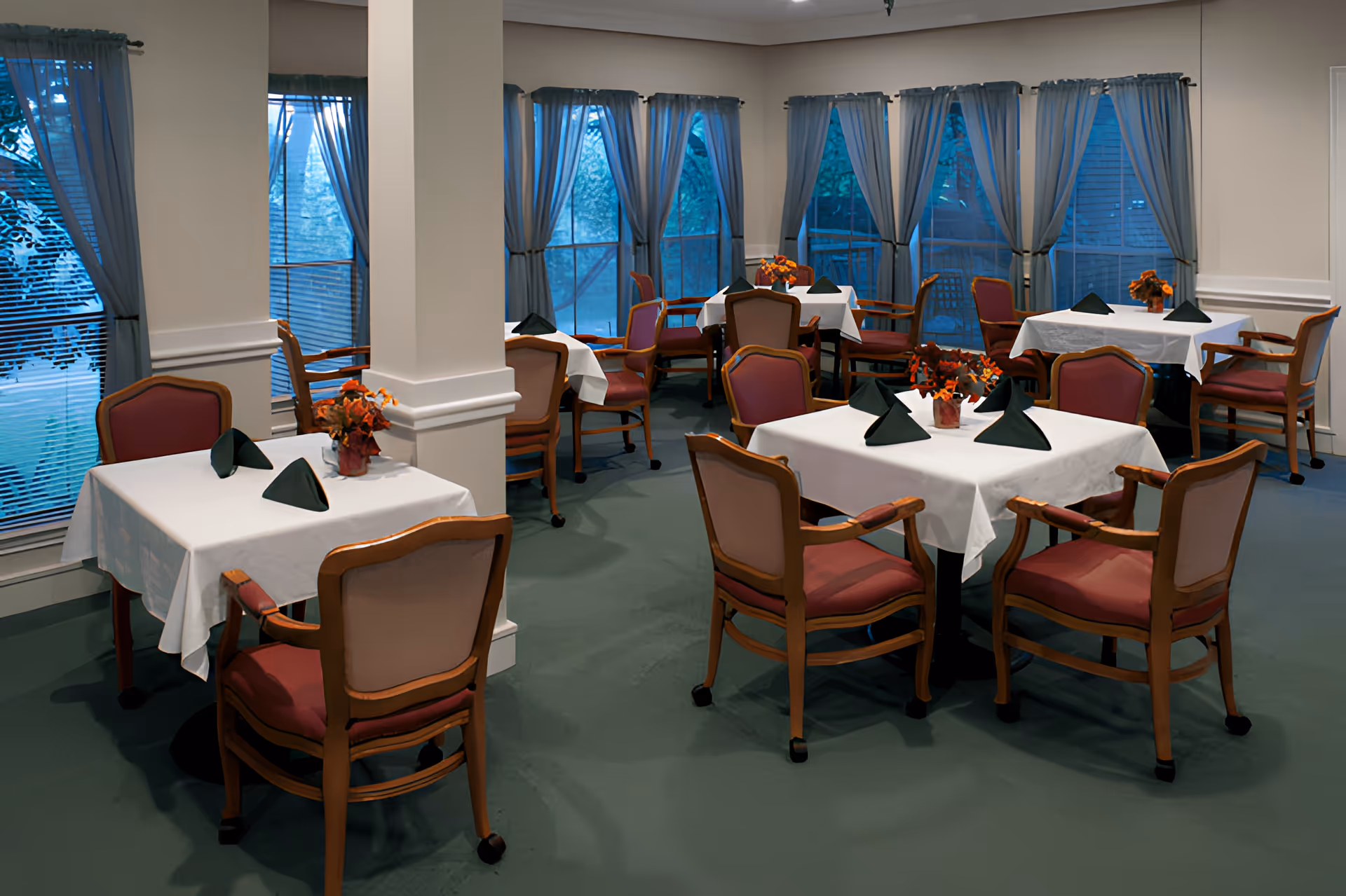 Dining room with multiple small tables set with white tablecloths, folded napkins, and floral centerpieces near large curtained windows.
