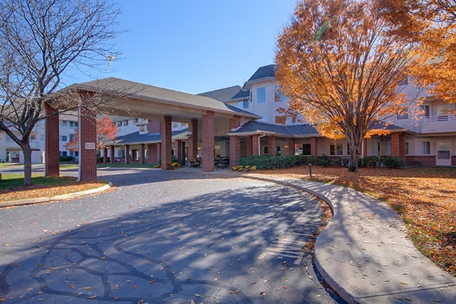 Exterior view of a senior living facility with a covered entrance supported by brick pillars, surrounded by trees with autumn foliage and a clear blue sky.