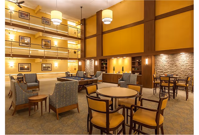 A spacious and well-lit common area in a senior living facility featuring multiple seating arrangements including round tables with yellow cushioned chairs, blue upholstered armchairs, and a pool table in the center. The room has warm yellow walls with wooden accents, stone wall panels, framed artwork, and modern pendant lights hanging from the ceiling.