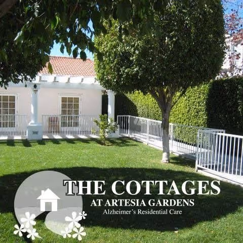 A well-maintained outdoor garden area at The Cottages at Artesia Gardens featuring green grass, a tree, white railings, and a building with white walls and a tiled roof in the background.