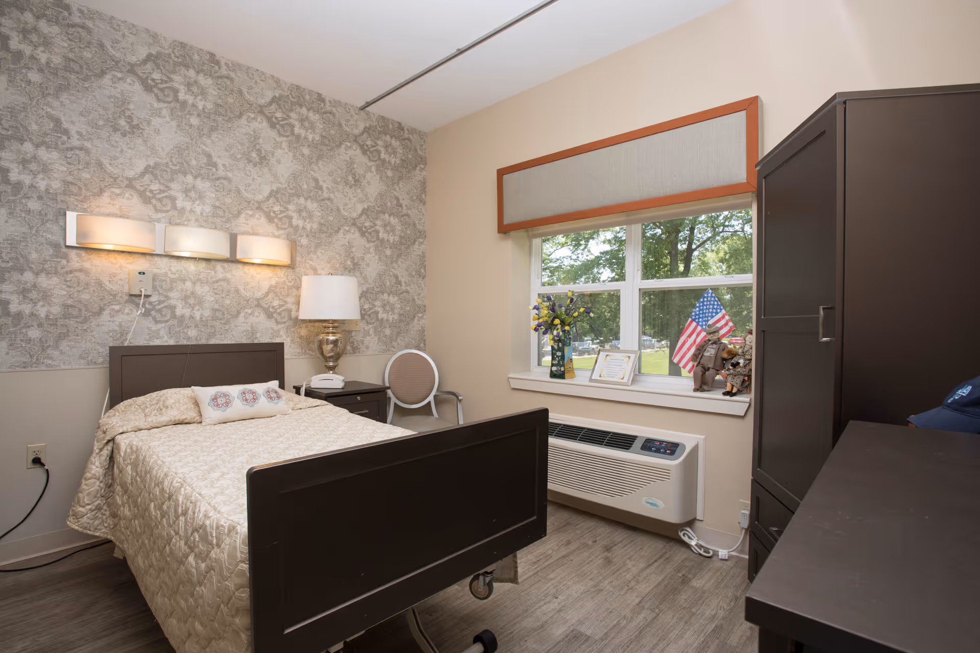 A neatly arranged bedroom in a senior living facility with a single bed covered in a beige quilt, a bedside table with a lamp and phone, a chair, a window with a view of trees outside, and a dark wooden wardrobe. The wall behind the bed has a patterned wallpaper and there are decorative items including a vase with flowers and small American flags on the windowsill.