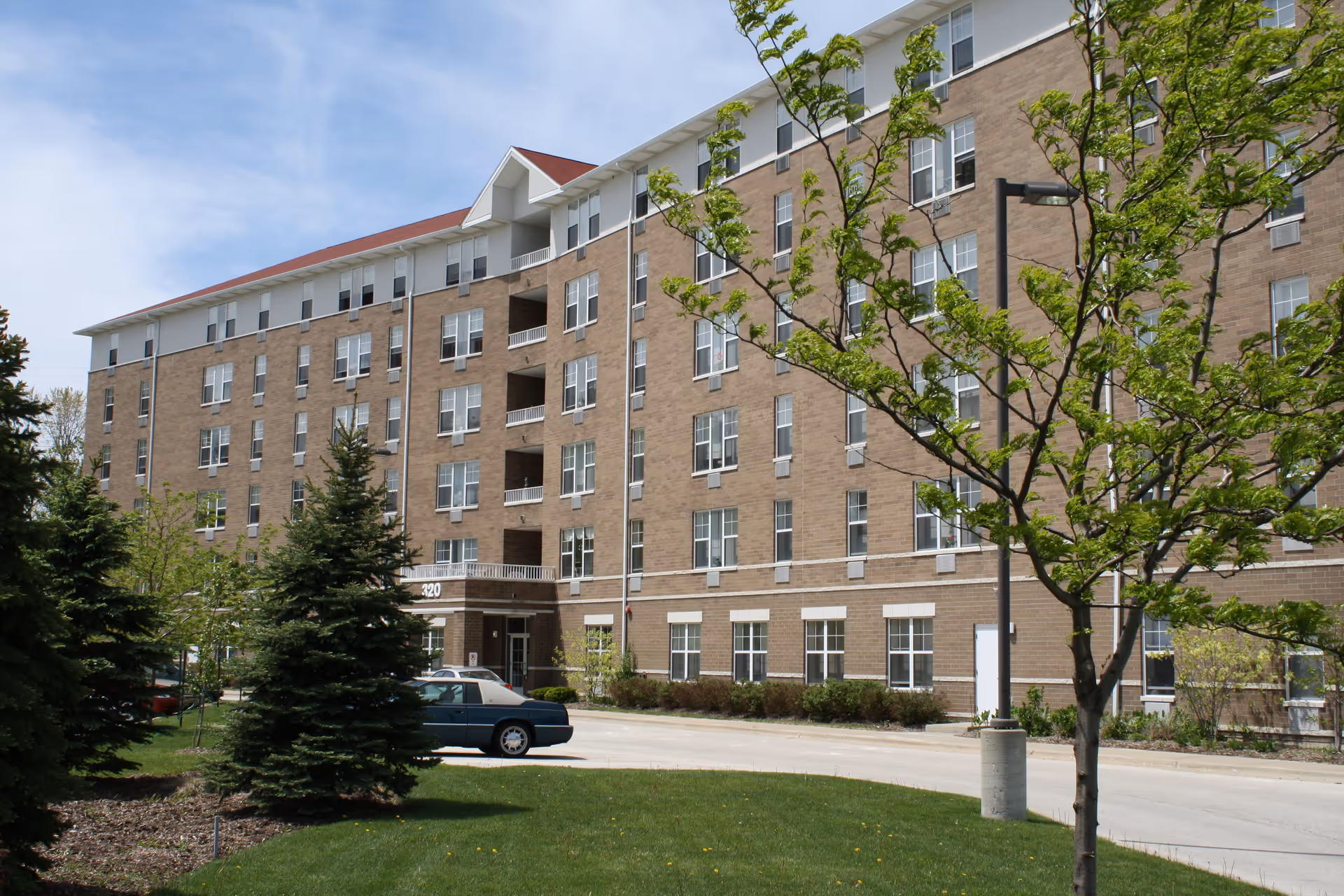 Exterior view of a multi-story brick apartment building with numerous windows, surrounded by green trees and a lawn under a partly cloudy sky. A car is parked near the entrance.
