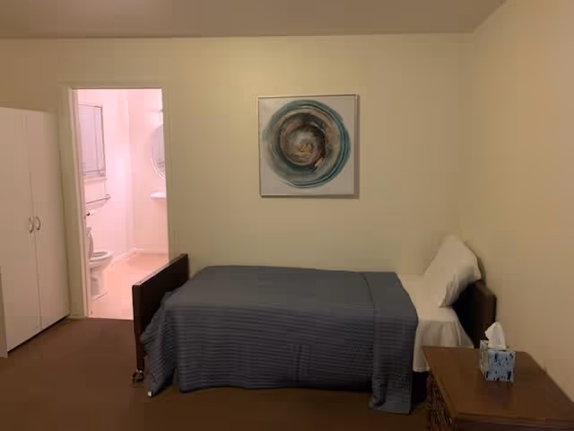 Single bed with a blue bedspread in a simple room with a nightstand, wardrobe, wall art, and an open door to a bathroom.