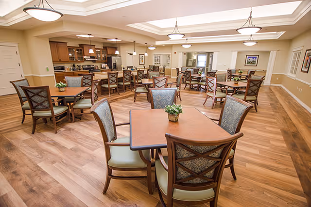 A spacious dining room with multiple wooden tables and cushioned chairs arranged neatly. The room features wood flooring, soft overhead lighting fixtures, and a kitchen area with cabinets and appliances in the background. Small potted plants are placed on the tables as centerpieces.