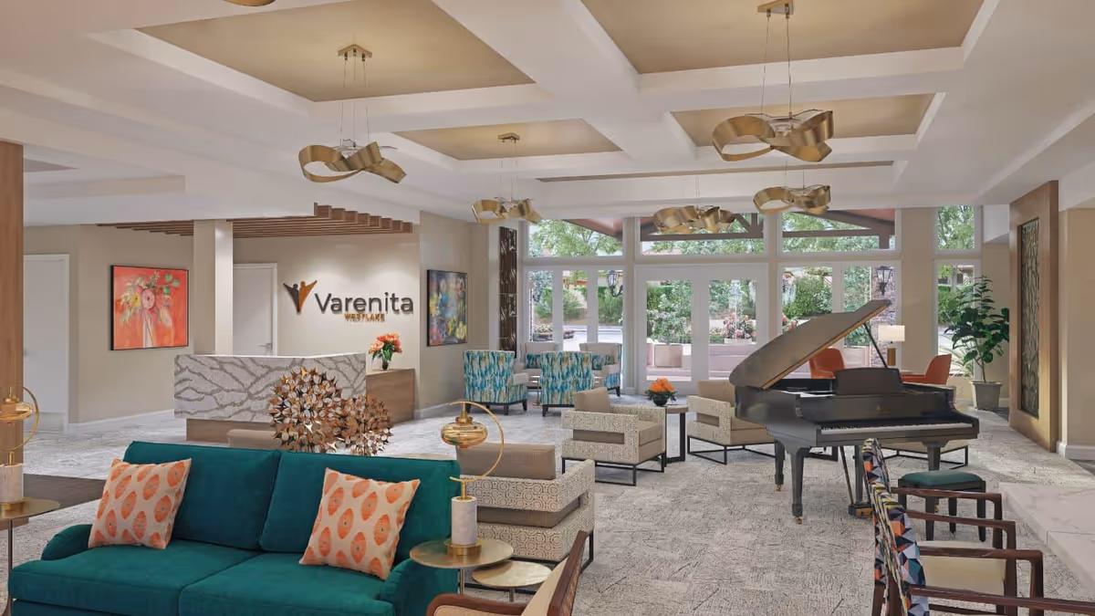 Bright senior living lobby with seating areas, a grand piano, and a reception desk bearing the Varenita logo in front of large windows.