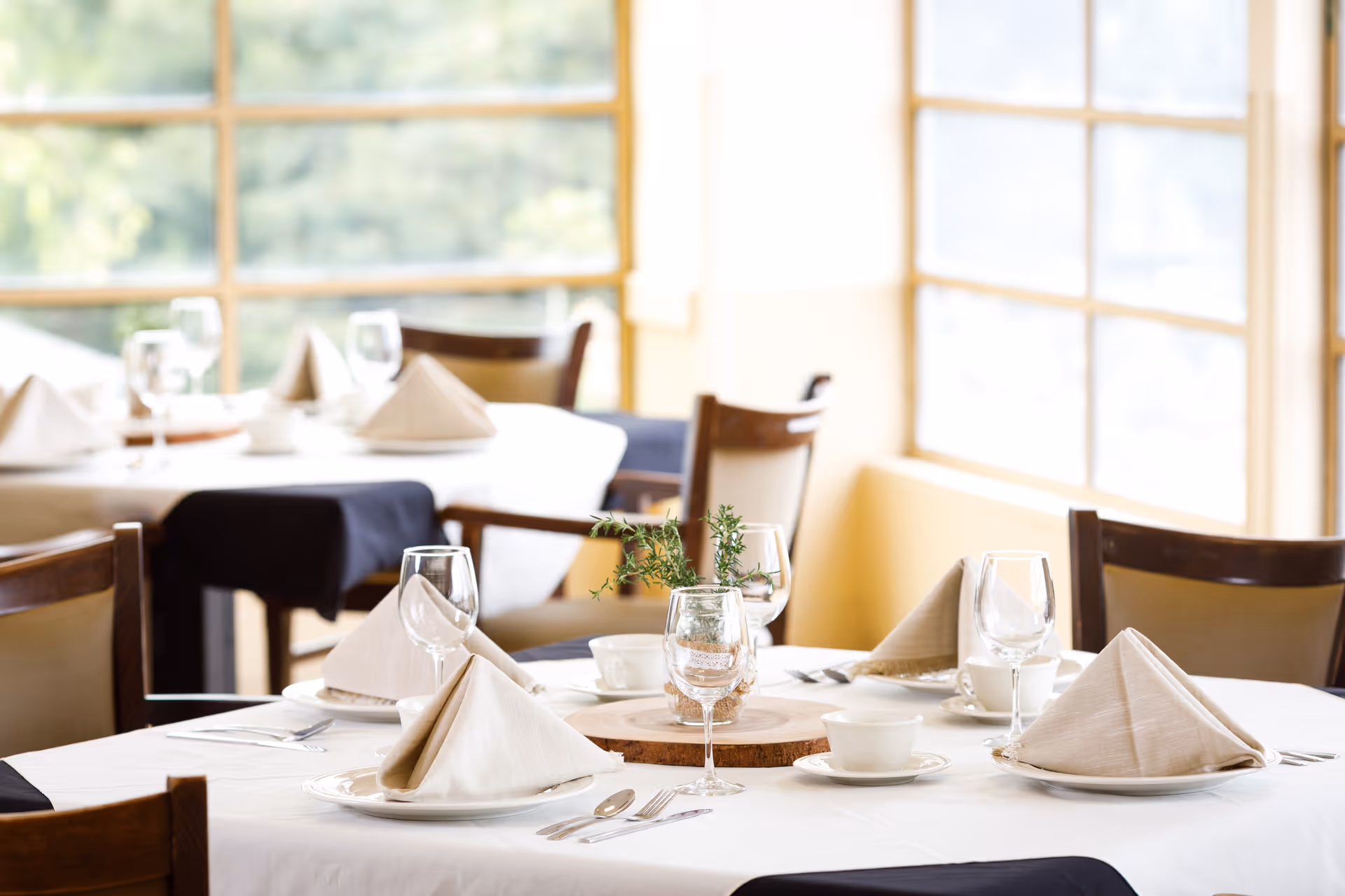 A bright dining room with tables set with folded napkins, glassware, cups, and small centerpieces near large windows.