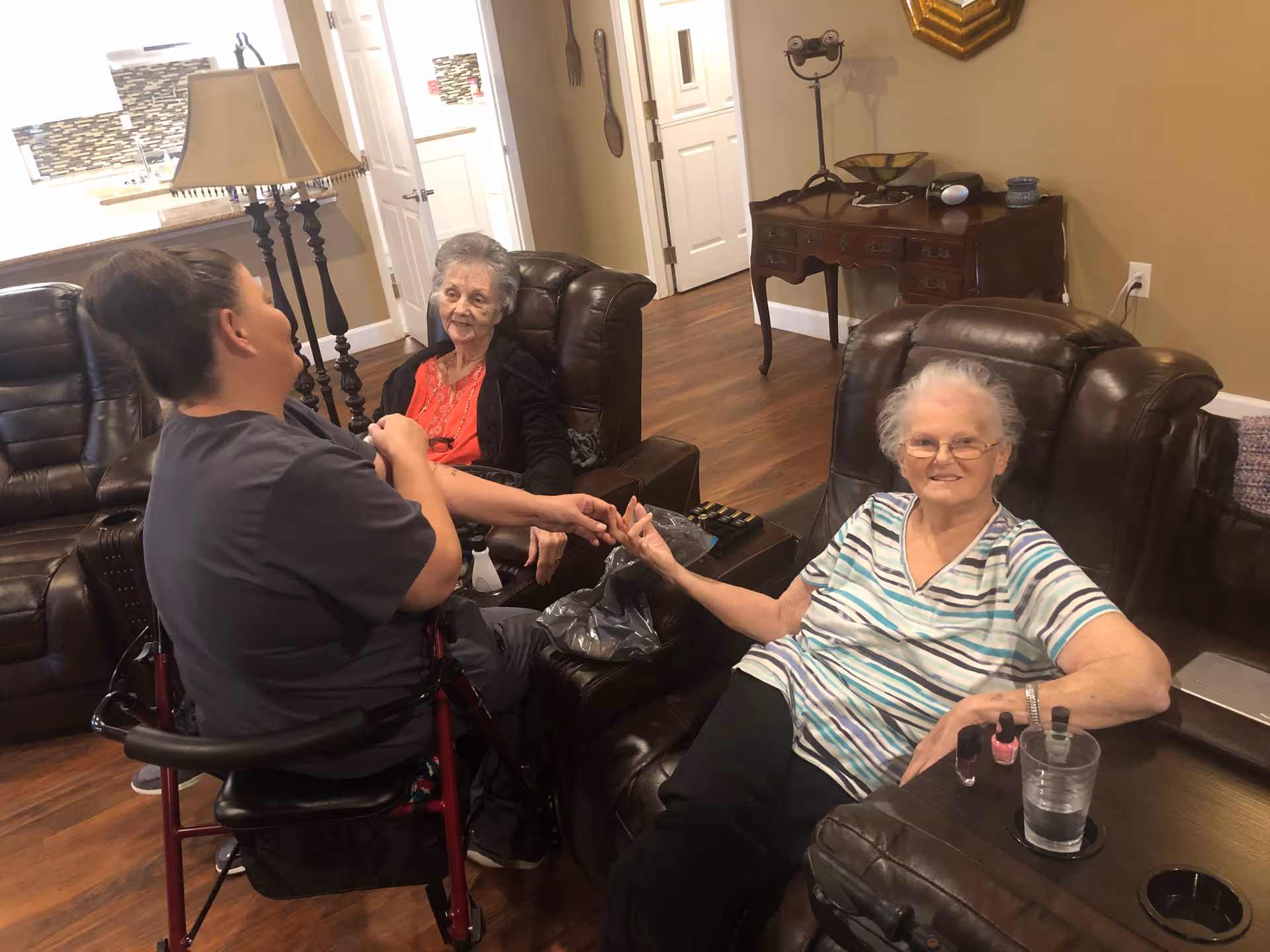 A caregiver is painting the nails of an elderly woman seated in a leather recliner, while another elderly woman sits nearby in a similar chair, smiling. The room has wooden floors, beige walls, and a wooden side table with decorative items. The background shows a kitchen area and a hallway with doors.