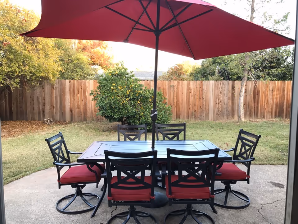 Patio dining table with six red-cushioned chairs and a large red umbrella on a concrete slab in a backyard with a wooden fence and trees.