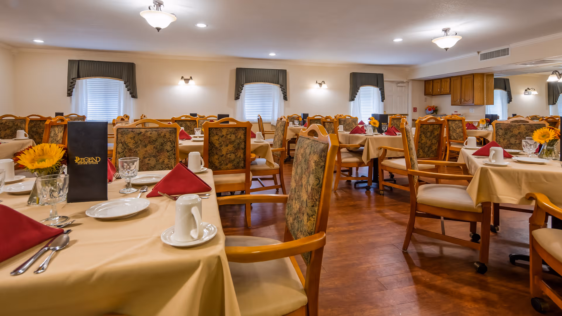 A dining room with multiple tables covered in beige tablecloths, each set with white plates, cups, silverware, and red folded napkins. The chairs have wooden frames with floral upholstered backs and seats. There are windows with green valances and wall-mounted lights. The room has wooden flooring and ceiling lights, with a small kitchen area visible in the background.