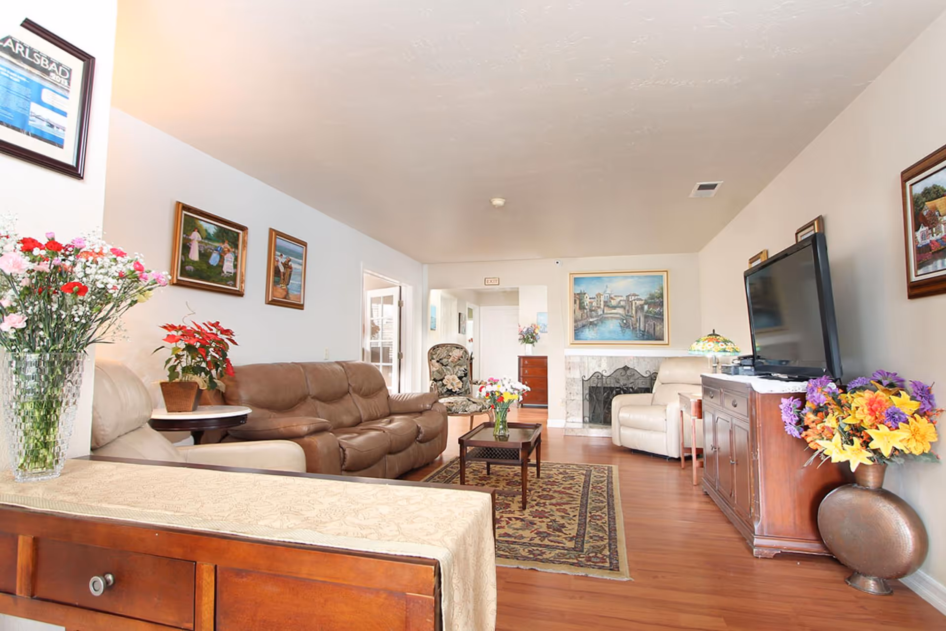A bright and cozy living room in Carlsbad Elder Care featuring a brown leather sofa, a beige recliner, a wooden coffee table with a vase of flowers, a large flat-screen TV on a wooden cabinet, and various framed paintings on the walls. The room has wooden flooring, a patterned area rug, and additional floral arrangements placed around the space.