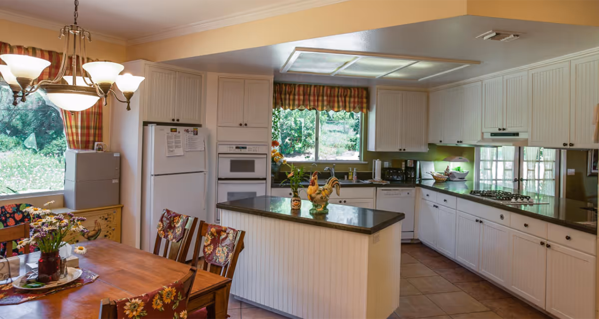 Bright country-style kitchen with a central island, white cabinets, granite countertops, and an adjacent dining table with floral chairs.